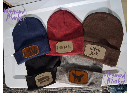 Set of four beanies with different text patches on a Wayward Market branded tray.